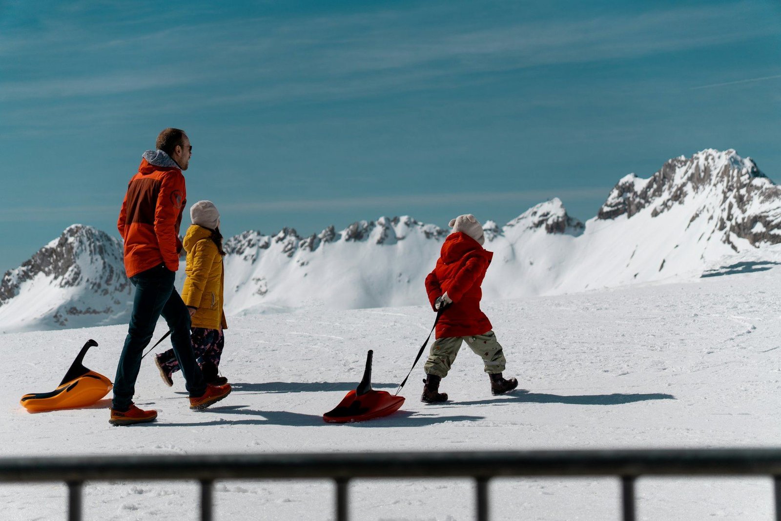 Three people in bright winter coats walking through deep snow and pulling sleds, with snowy peaks behind them