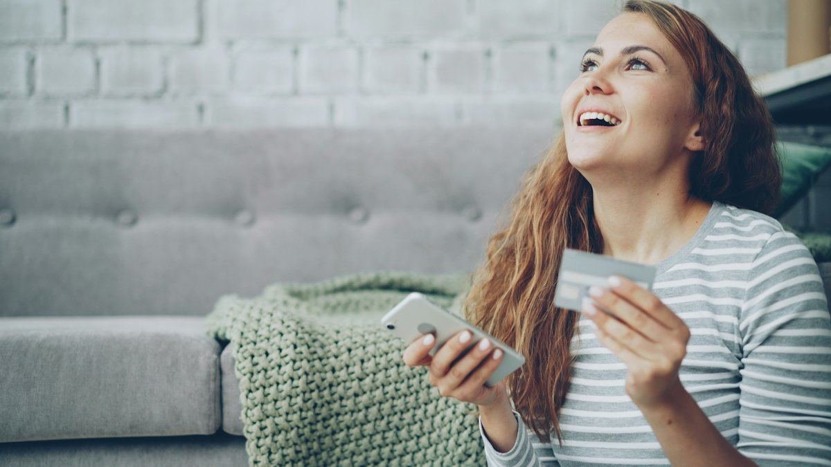 Une femme souriante sur un canapé, une carte bancaire dans une main et son téléphone dans l’autre